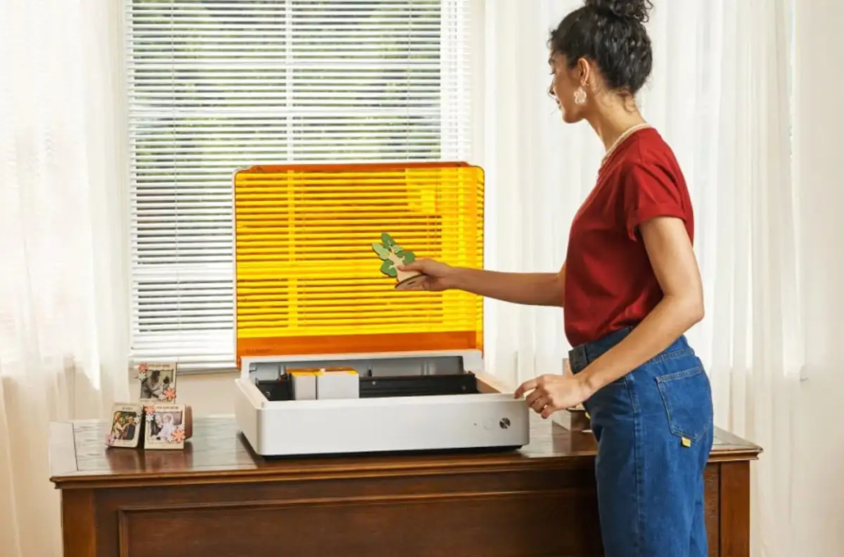 Frau in rotem T-Shirt und blauer Jeans bedient den xTool M1 Ultra Lasergravierer mit geöffnetem gelbem Schutzdeckel auf einem Holztisch